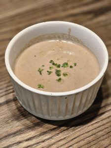 A bowl of cream of Mushroom Bisque soup, garnished with fresh parsley and a drizzle of oil, set against a wooden surface. The soup's smooth, light brown color and the bowl's simple, ridged design create a clean and inviting presentation. It was captured in Kozhikode, Kerala.
