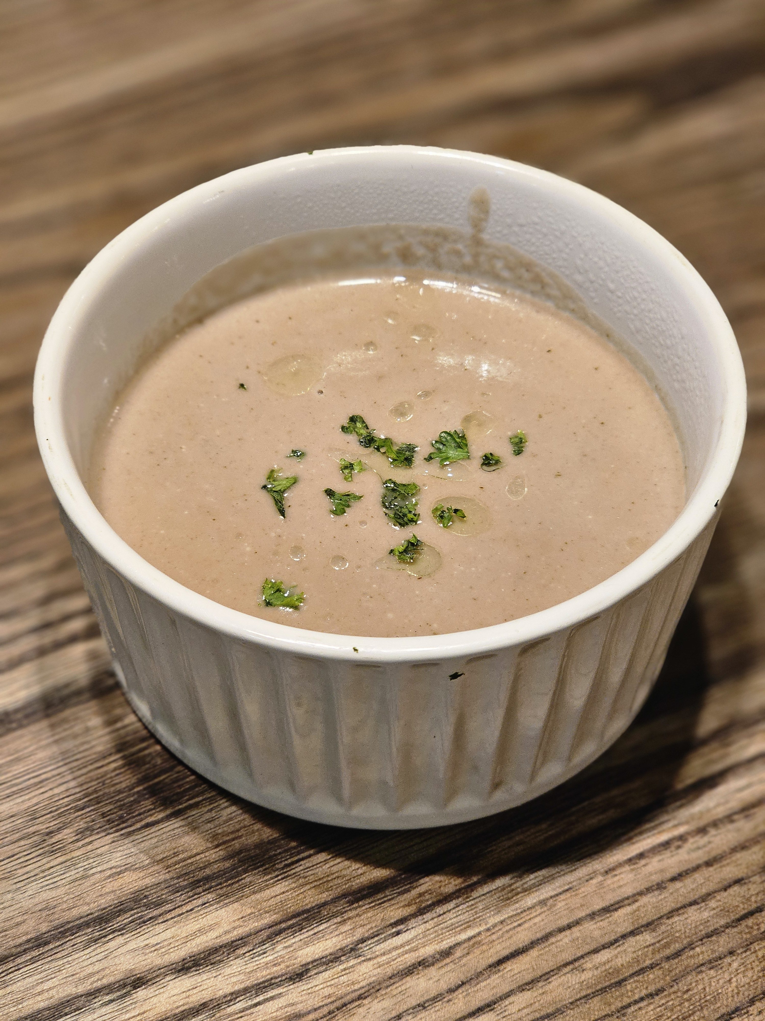 A bowl of cream of Mushroom Bisque soup, garnished with fresh parsley and a drizzle of oil, set against a wooden surface. The soup's smooth, light brown color and the bowl's simple, ridged design create a clean and inviting presentation. It was captured in Kozhikode, Kerala.