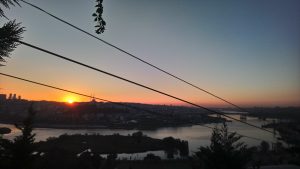A serene sunrise over a cityscape, with the sun rising above the horizon and casting a warm golden glow. In the foreground, silhouettes of trees and power lines frame the scene, while a river reflects the morning sky.