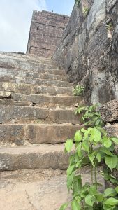 Stone steps lead up to a weathered, ancient brick tower with small rectangular windows. The steps are flanked by a rocky wall on the right, with small green plants growing among the stones and at the base. The sky above is clear with a few clouds.