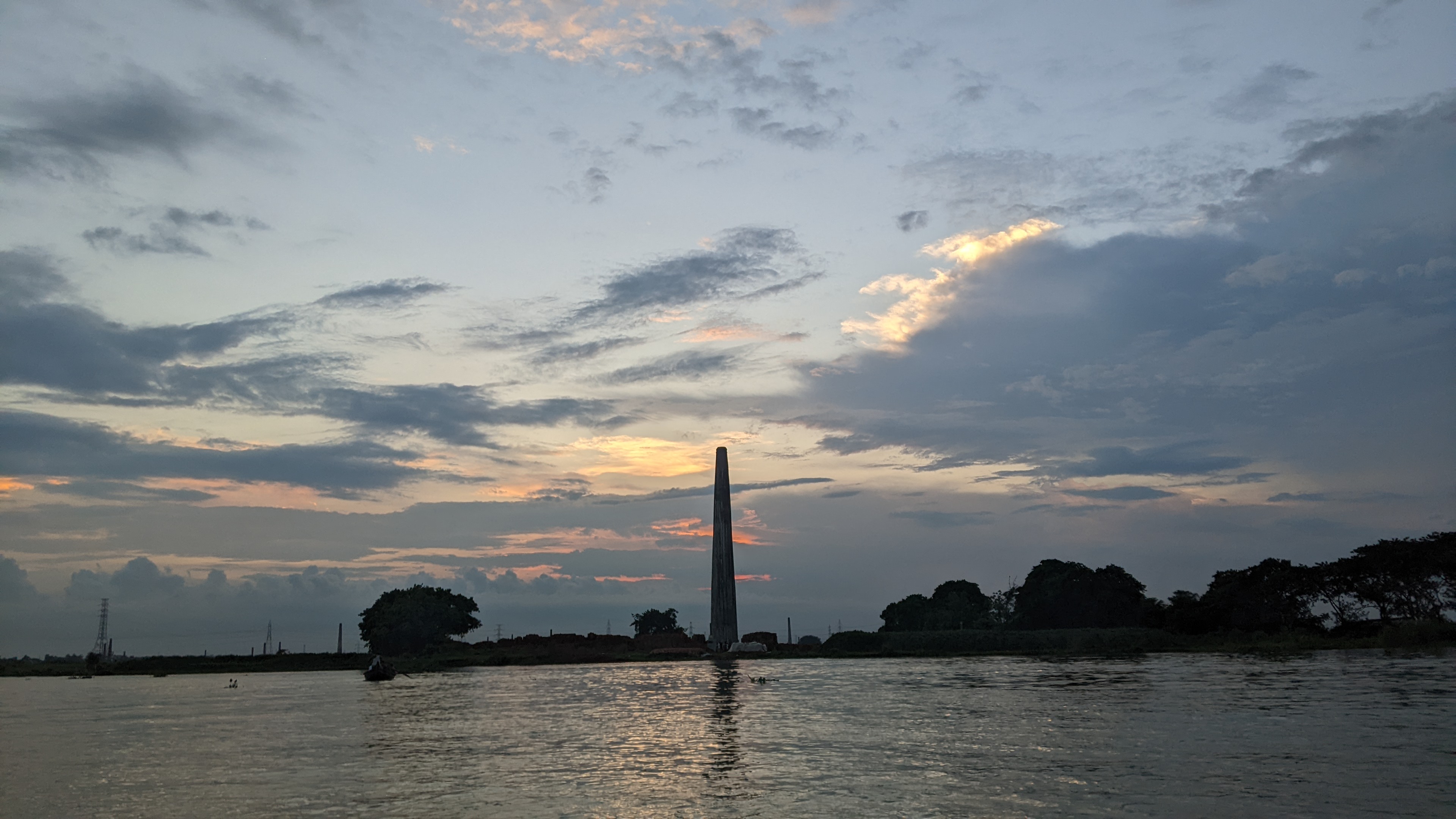 A tall, narrow chimney stands silhouetted against a cloudy sky at sunset, with trees and a body of water in the foreground.