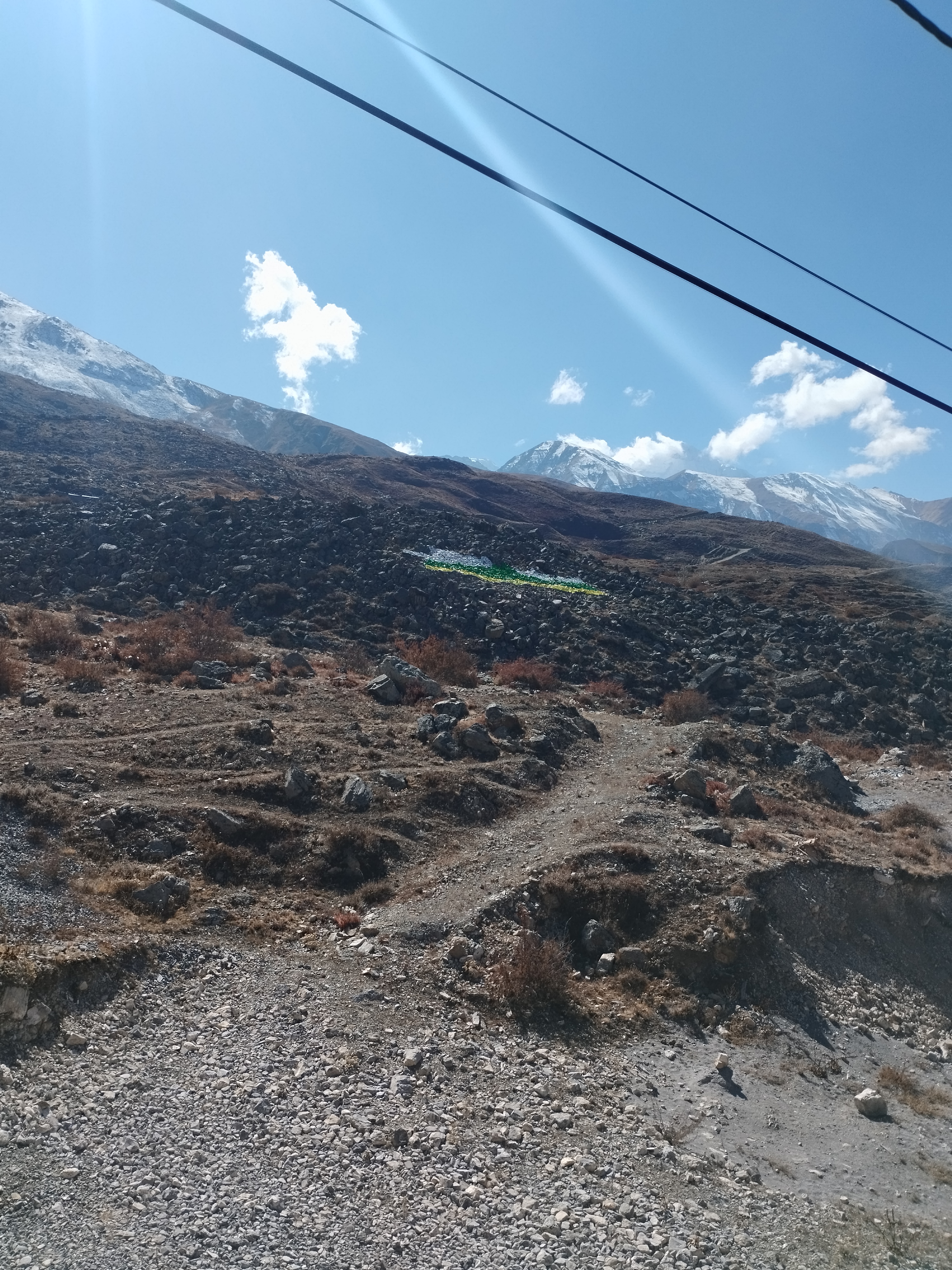 Rocky landscape with a dirt path, barren terrain, and sparse vegetation, set against a backdrop of snow-covered mountains under a clear blue sky with a few clouds.