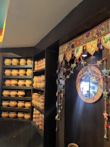 A wooden shelving unit filled with numerous clay pots is situated on the left side. On the right, a decorative mirror with a colorful beaded and tassel design hangs amidst vibrant fabric with intricate patterns. The scene is part of a warmly lit, traditional-style interior.