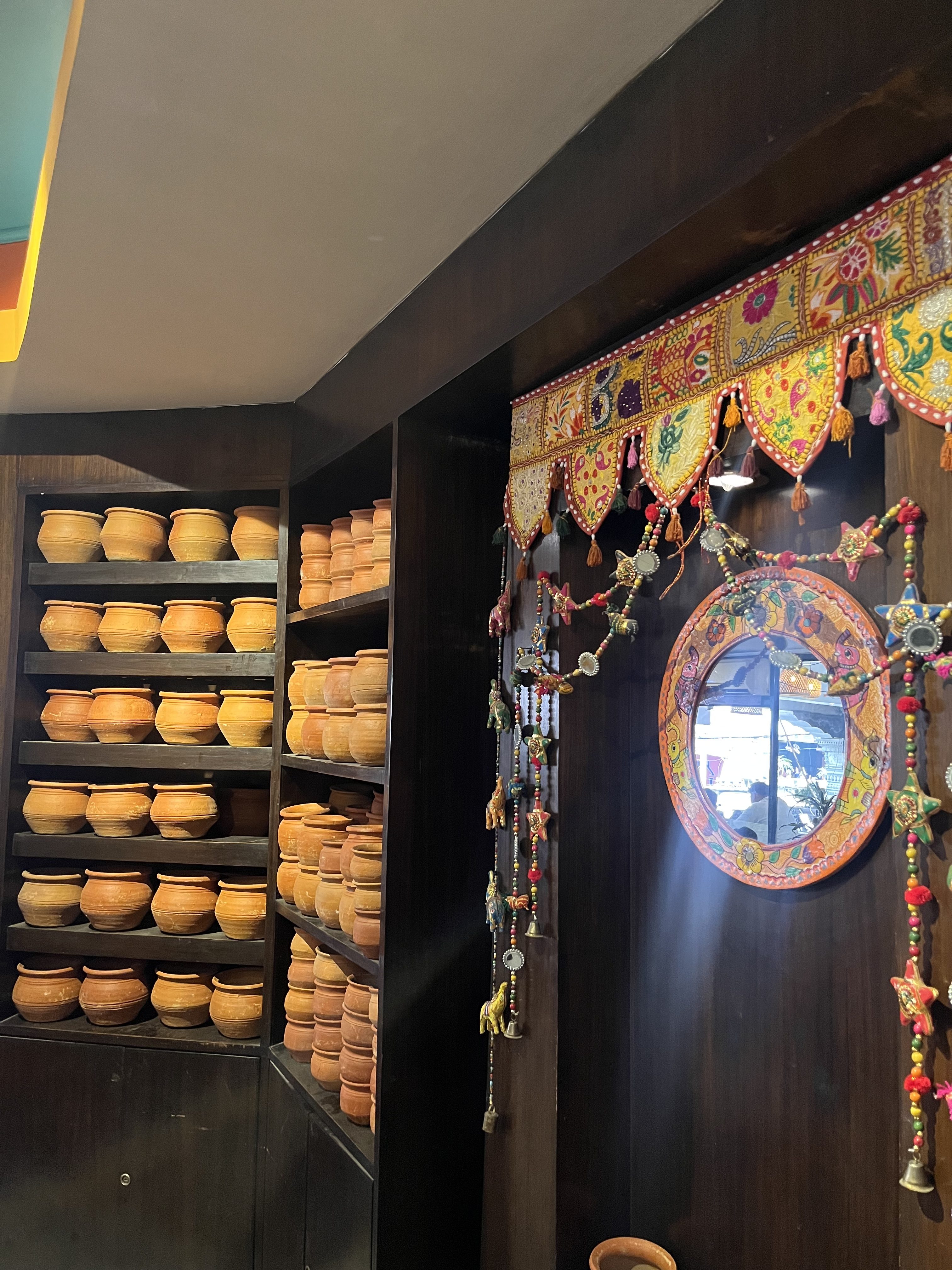 A wooden shelving unit filled with numerous clay pots is situated on the left side. On the right, a decorative mirror with a colorful beaded and tassel design hangs amidst vibrant fabric with intricate patterns. The scene is part of a warmly lit, traditional-style interior.