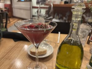 Closeup of a pink cocktail in a martini glass, rosemary and cranberries in the drink.  Next to it is the top of an olive oil carafe.