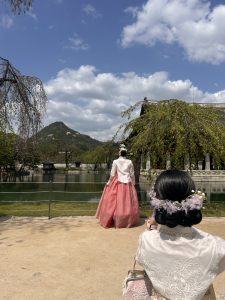 A serene outdoor scene featuring two women in traditional Korean hanbok. One woman, seen from behind, wears a pink skirt and a white top, standing near a calm body of water with a picturesque mountainous backdrop.