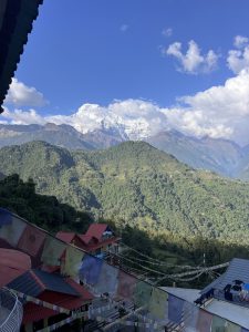 A scenic view of a mountainous landscape with lush green hills in the foreground and snow-capped peaks in the background. There are colorful prayer flags strung across the scene, and rooftops of buildings can be seen with red and dark-colored tiles. The sky is clear with a few scattered clouds.