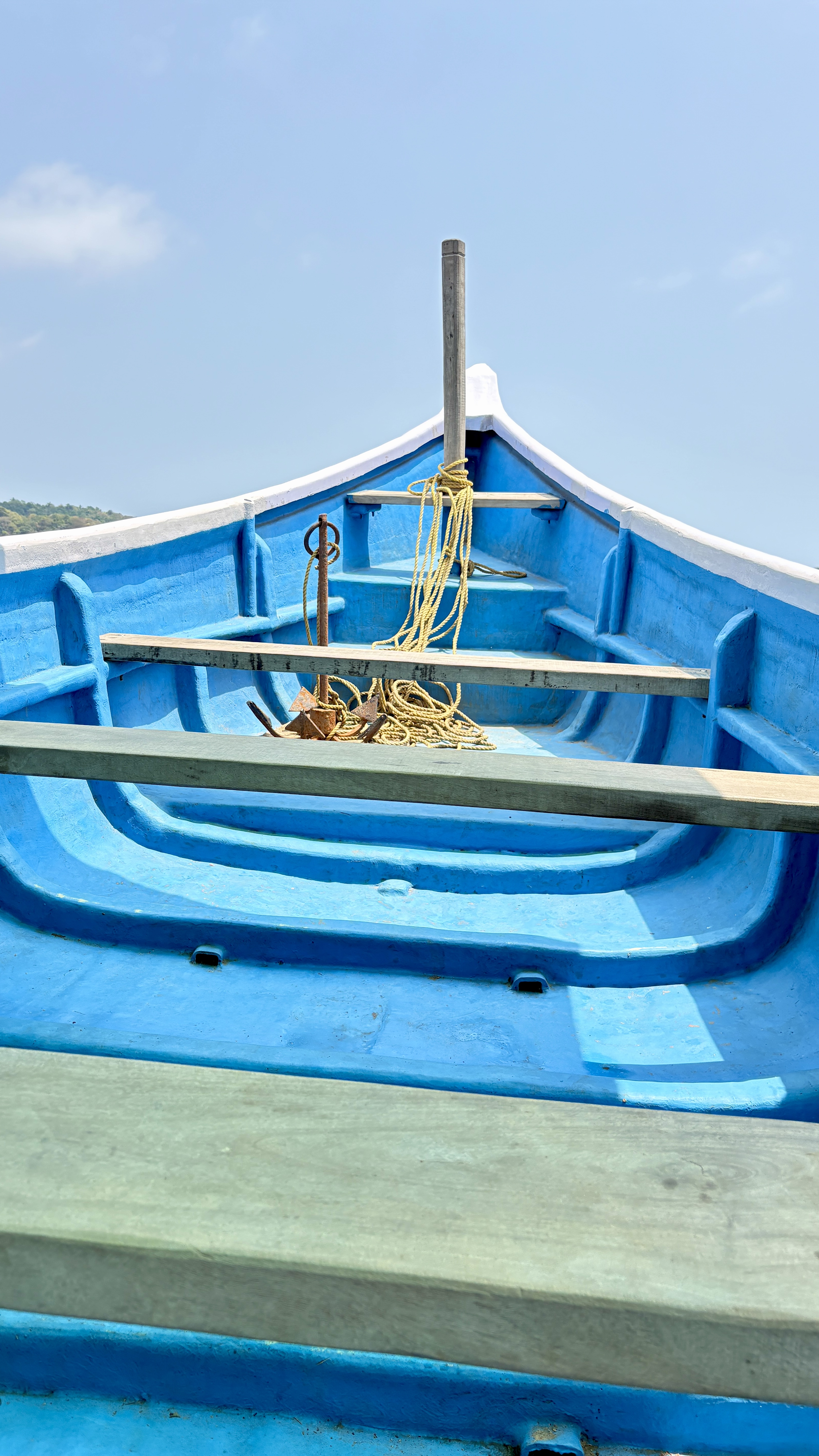 Interior view of a bright blue wooden boat with wooden planks as seats and coiled rope near the anchor at the front. The sky is clear and blue, with a hint of land visible in the background.