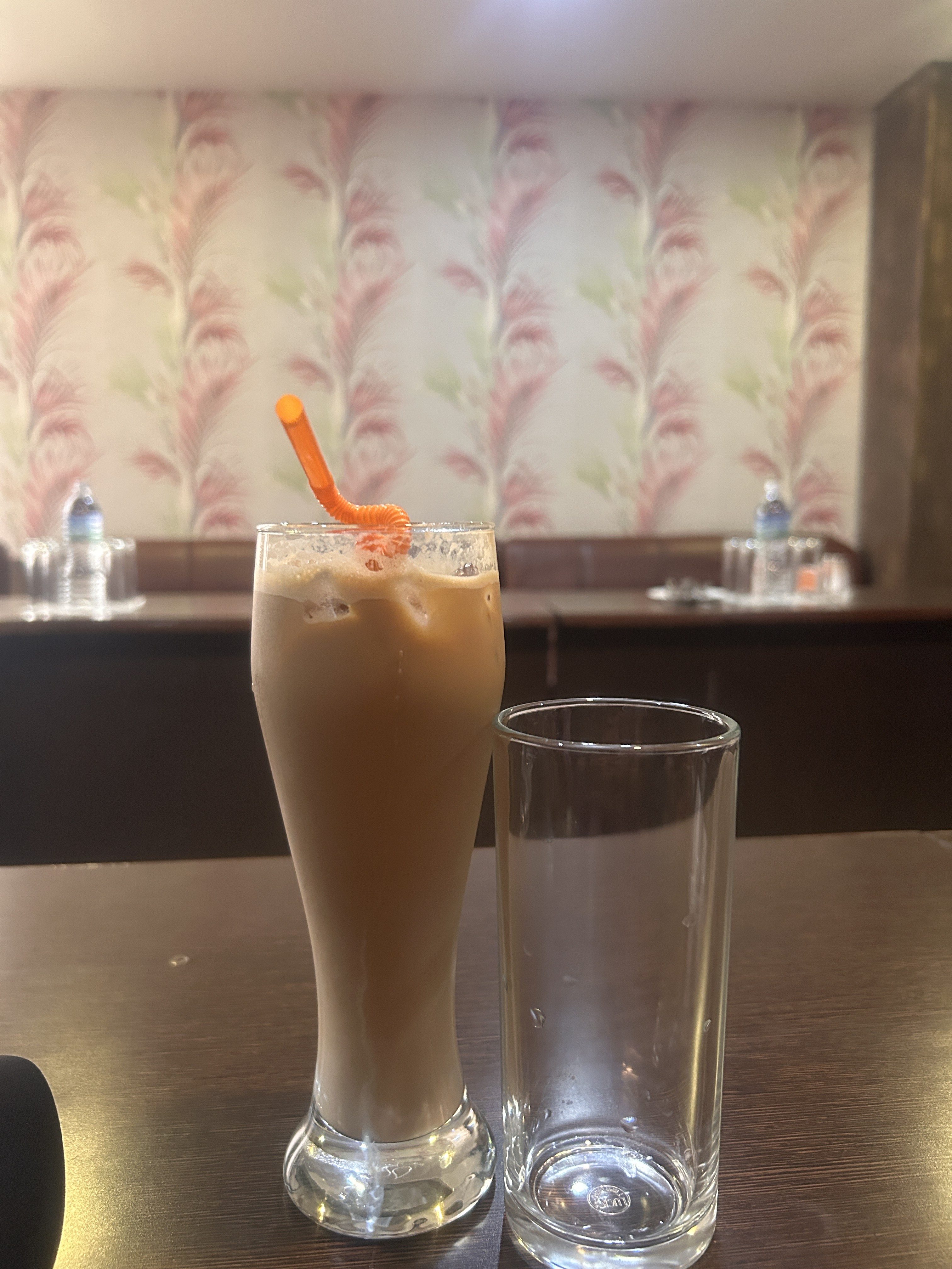 A tall glass filled with an iced coffee drink and an orange straw next to an empty glass on a table