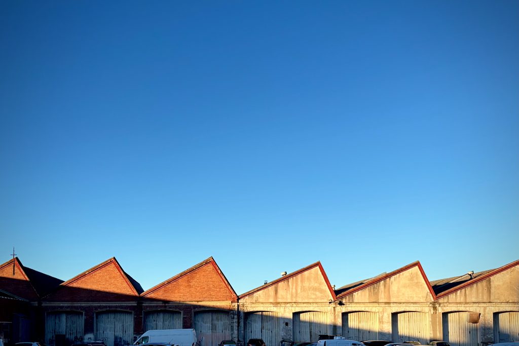 A row of industrial warehouse buildings with red triangular roofs against a clear blue sky. Several vehicles are parked in front of the warehouses.