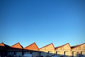 A row of industrial warehouse buildings with red triangular roofs against a clear blue sky. Several vehicles are parked in front of the warehouses.

