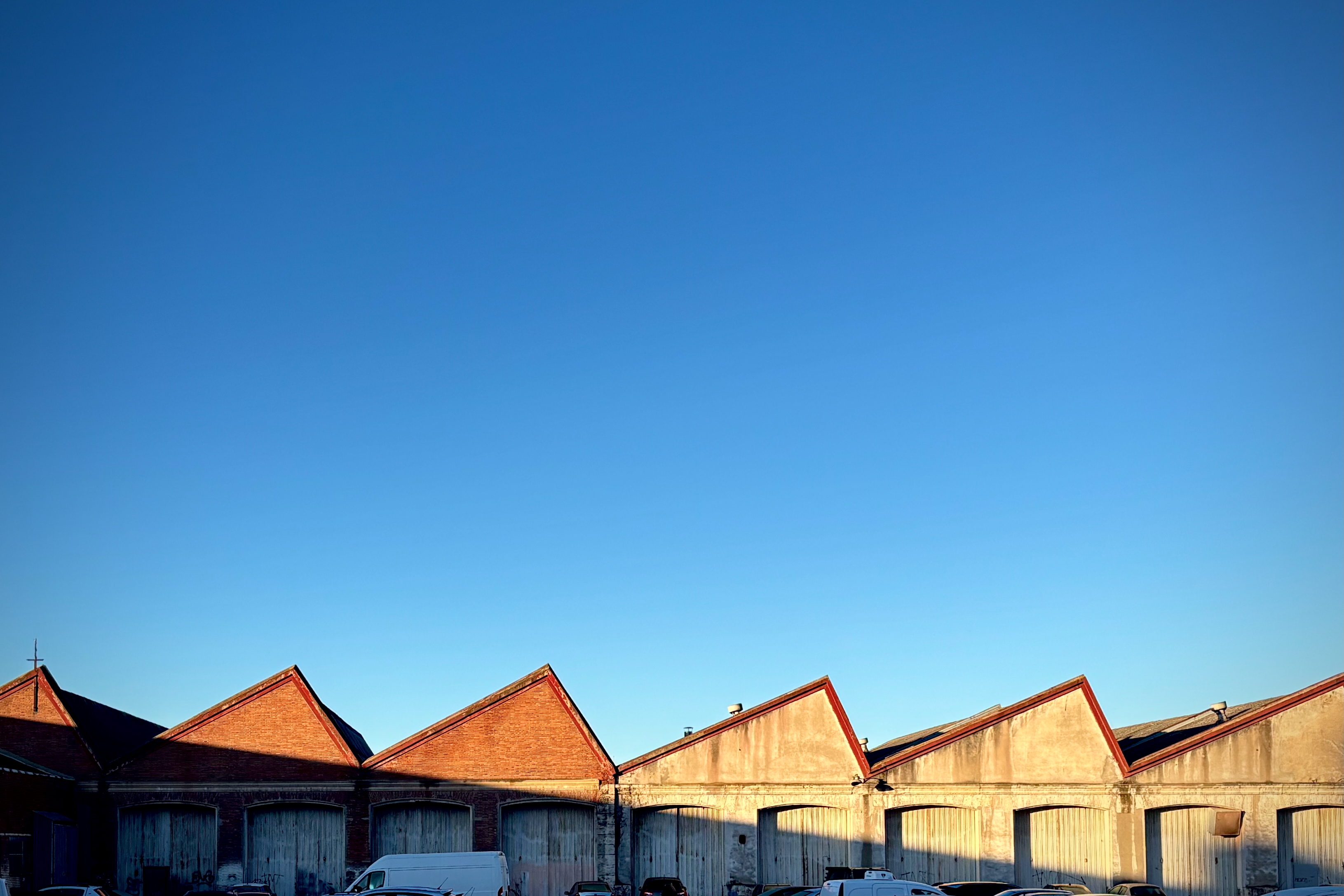 A row of industrial warehouse buildings with red triangular roofs against a clear blue sky. Several vehicles are parked in front of the warehouses.
