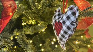Closeup of an ornament made of cloth, shaped like a heart, in the branches of an evergreen tree with lights. A bit of red ribbon is visible on the left.
