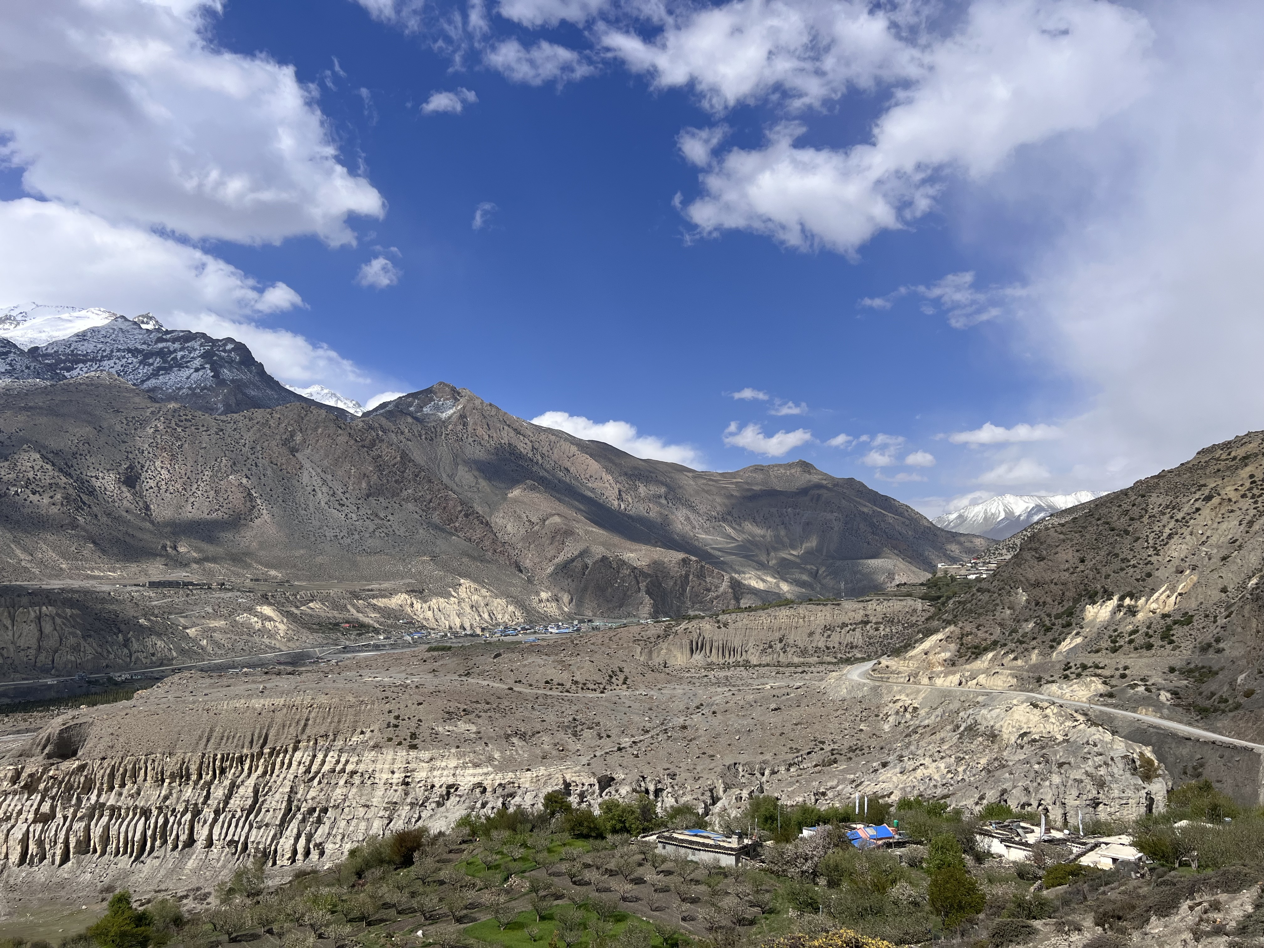 A mountainous landscape of rocky terrain and patches with rugged peaks under a partly cloudy blue sky. Snow-capped mountains can be seen in the background.