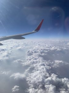A view from an airplane window showing a wing with a red tip against a clear blue sky filled with fluffy white clouds below. In the distance, a contrail can be seen from another aircraft. The sunlight creates a shiny effect, illuminating the scene.