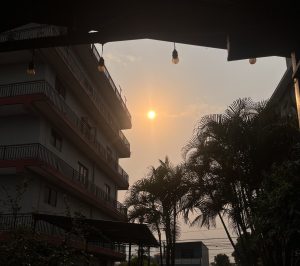 A sunset is visible behind a multi-story building, partially obscured by trees. 