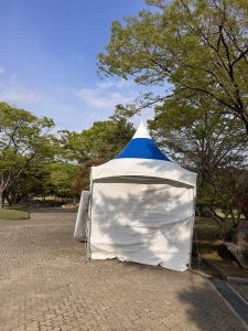 A white tent with a blue top is set up in a park, partially obscured by trees. 