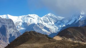 A scenic view of snow-capped mountains under a clear blue sky, with rugged terrain in the foreground and a few small figures visible on a hill.
