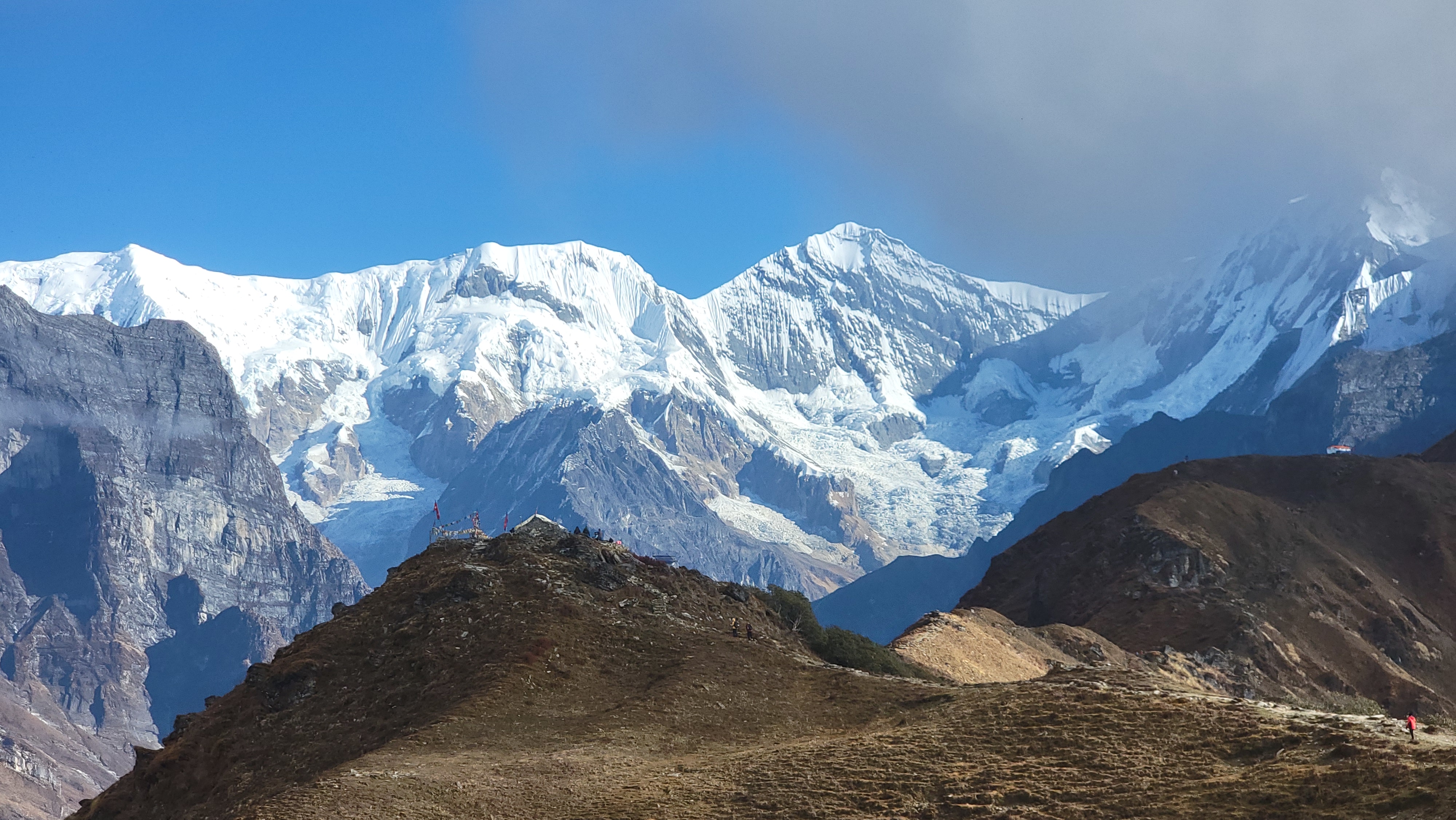 A scenic view of snow-capped mountains under a clear blue sky, with rugged terrain in the foreground and a few small figures visible on a hill.