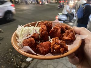 A clay plate held in a hand containing fried chicken pieces and shredded cabbage, with a street scene in the background.