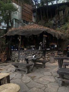A rustic outdoor bar with a stone facade and a thatched roof covered in leaves, surrounded by trees and corrugated metal sheets. Wooden tables and benches are placed on a cobblestone floor, and various bottles and decorations are visible on the bar counter.