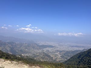 A panoramic view of a hilly landscape with green valleys and a city nestled in between. In the distance, majestic snow-capped mountains are visible under a clear blue sky.

