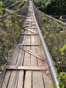 A weathered wooden suspension bridge stretches across an area surrounded by greenery.