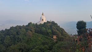 A white stupa with a golden top is situated on a lush green hill, surrounded by dense trees. The background features a hazy view of distant mountains and a cityscape, under a clear blue sky.