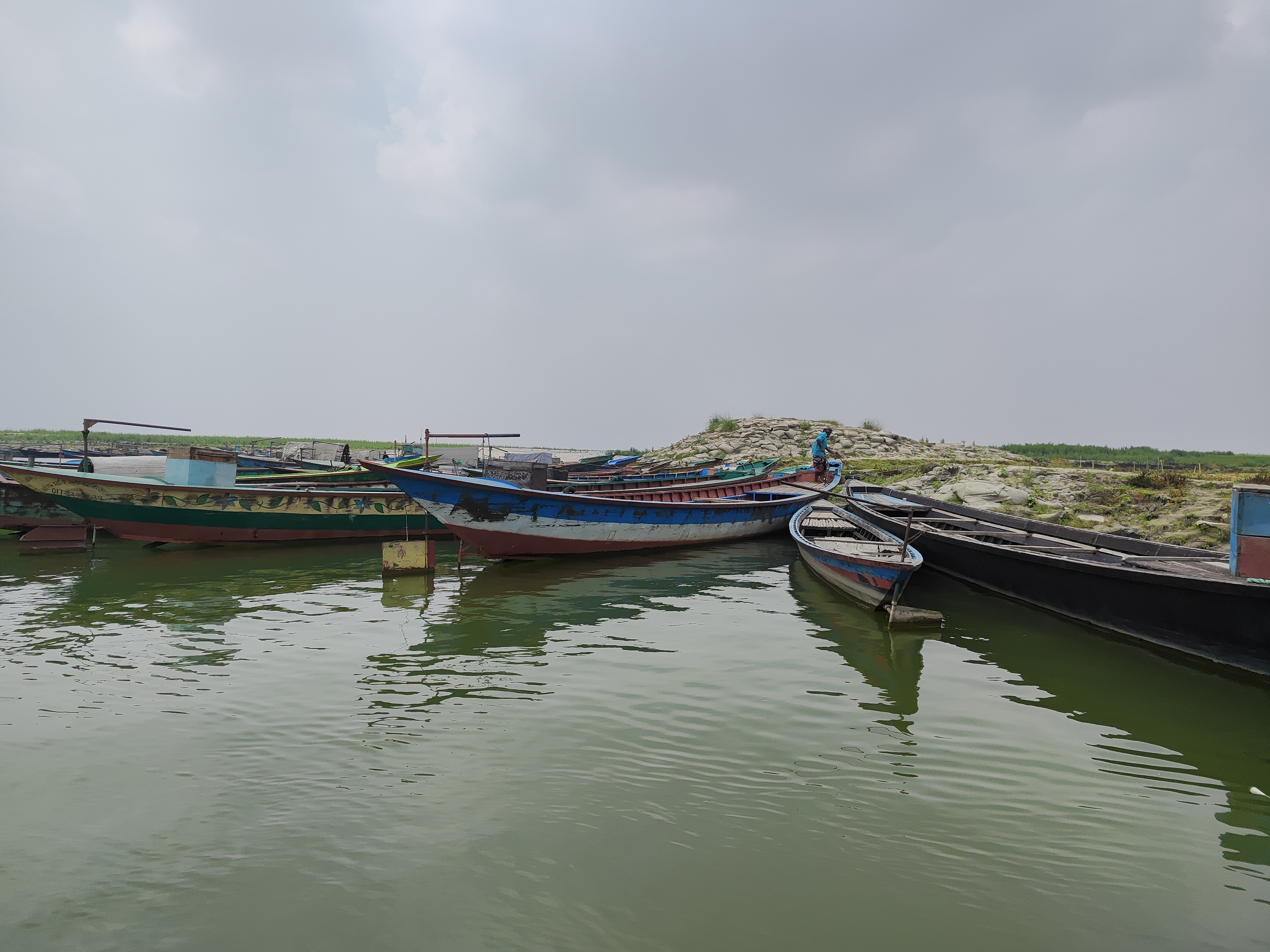 Several wooden fishing boats are moored on calm greenish water near a rocky, grassy shoreline under a cloudy sky. A person stands on one of the boats near the shore.