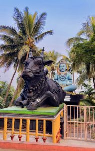 Large statue of Nandi the sacred bull with Lord Shiva seated behind it, set against a backdrop of tall palm trees and a clear blue sky, enclosed by a yellow and red fence.