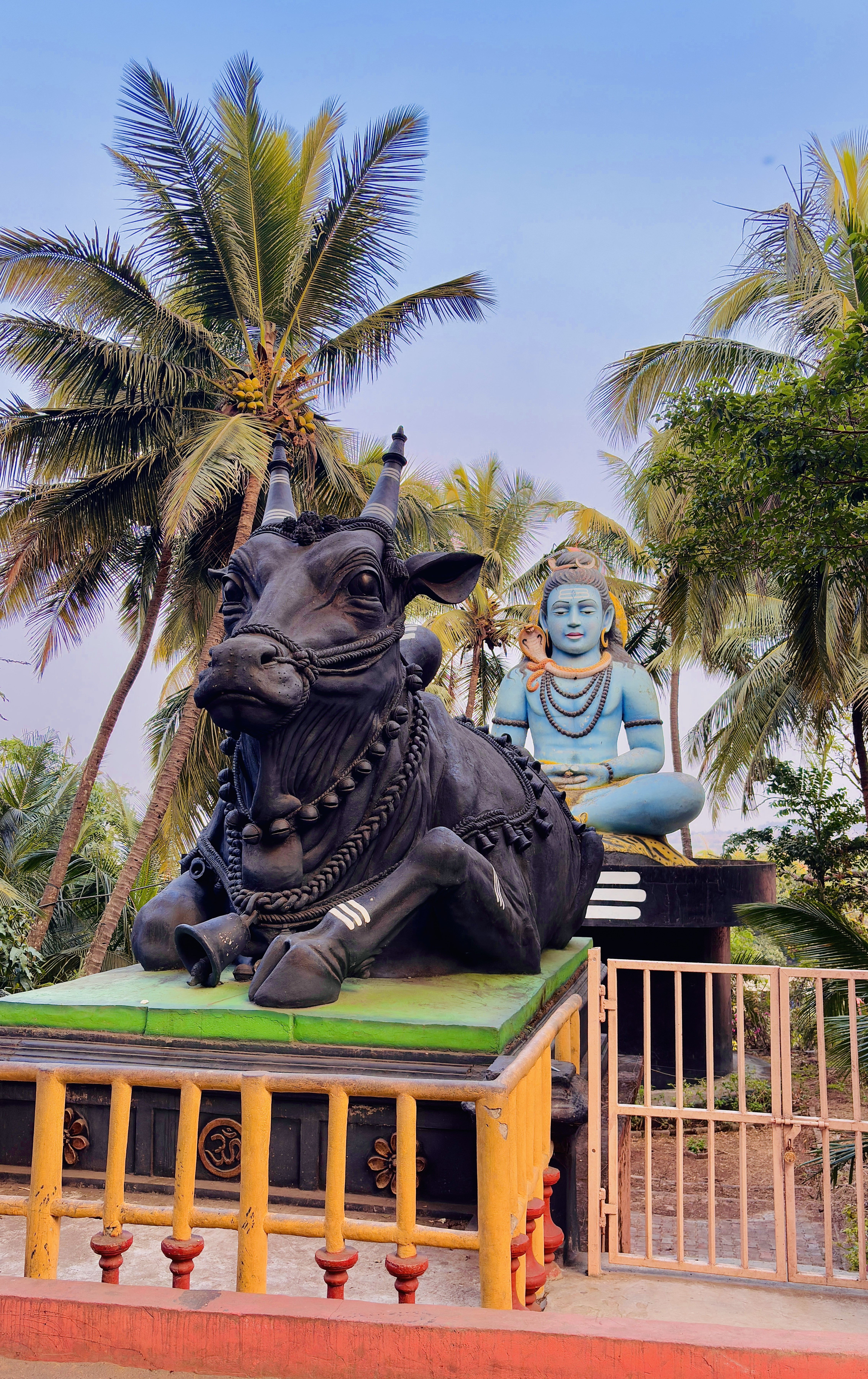 Large statue of Nandi the sacred bull with Lord Shiva seated behind it, set against a backdrop of tall palm trees and a clear blue sky, enclosed by a yellow and red fence.