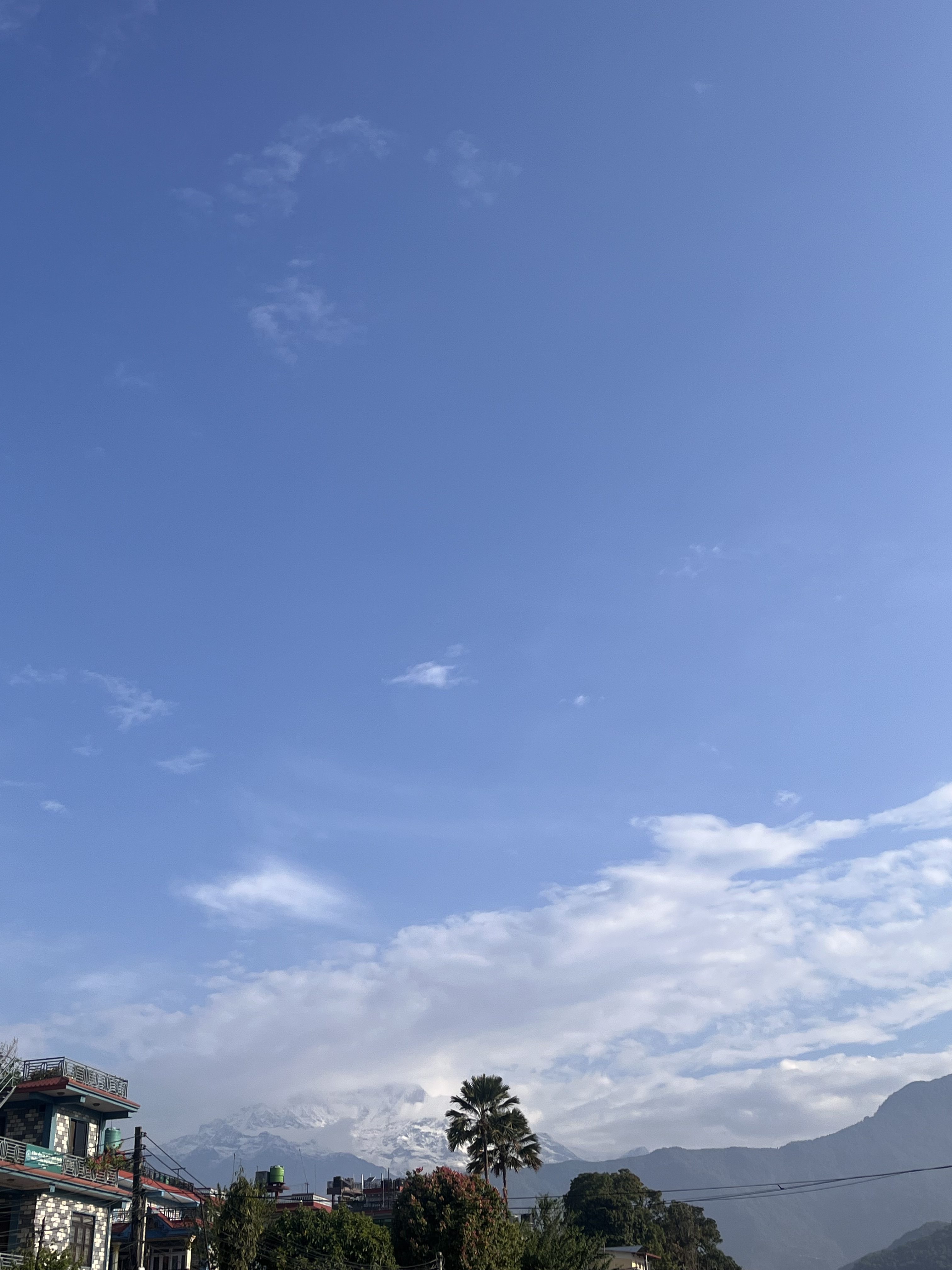 A serene landscape featuring a clear blue sky with scattered clouds above a mountainous region. Below, buildings with rooftops and lush green trees are visible, with snow-capped mountains in the background.