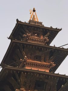 A close-up view of an intricately designed, multi-tiered pagoda-style temple roof with ornate carvings and a golden trident at the pinnacle, under a clear sky.