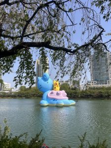 A large blue inflatable sculpture of a cat-like creature with a small yellow character sitting on its back is floating on a calm lake, surrounded by trees. In the background, modern buildings can be seen against a clear blue sky