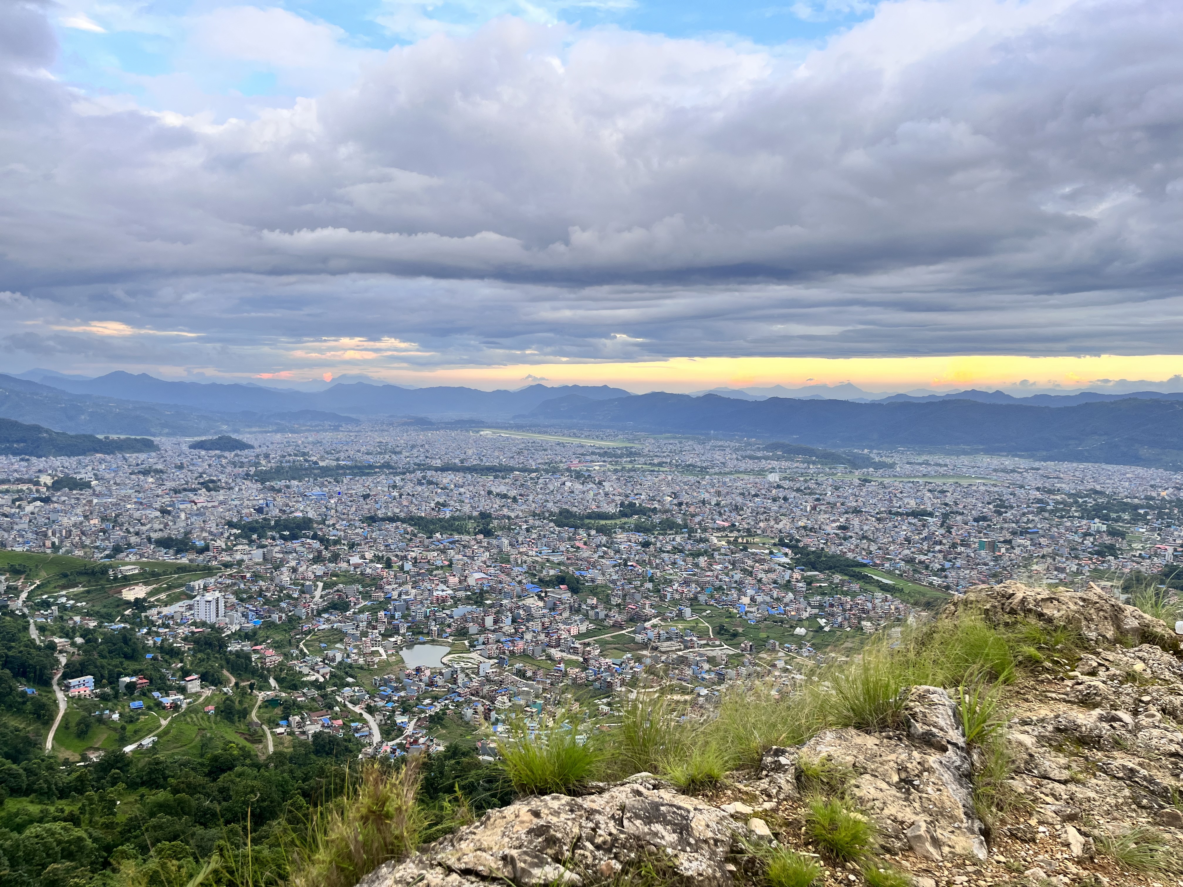 A panoramic view of a cityscape with numerous buildings, surrounded by lush greenery and mountains in the distance. The sky is partially cloudy with patches of blue and a faint yellow-orange hue near the horizon, suggesting a sunset or sunrise. In the foreground, there are rocky and grassy terrain.