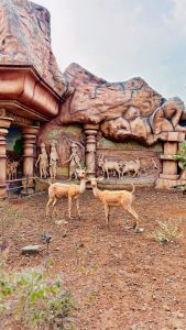 Sculpted diorama depicting rural village life with farmers, oxen, and trees carved into a rock-style backdrop, with two life-sized deer statues standing in the foreground on reddish soil.