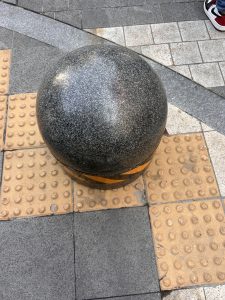 A large, smooth, dark granite sphere sits on a patterned pavement, surrounded by square tiles of varying colors, including gray and beige. 