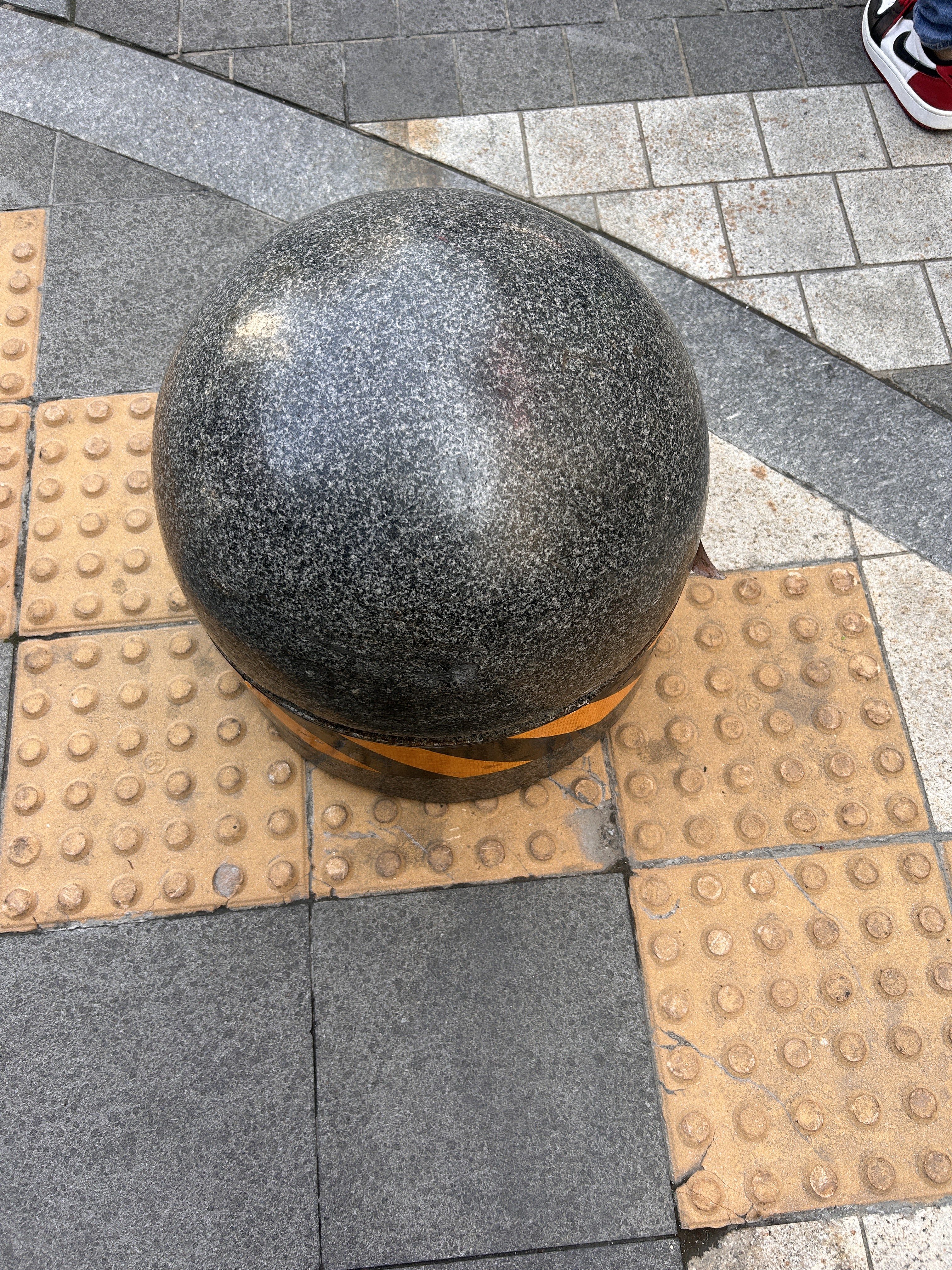 A large, smooth, dark granite sphere sits on a patterned pavement, surrounded by square tiles of varying colors, including gray and beige.