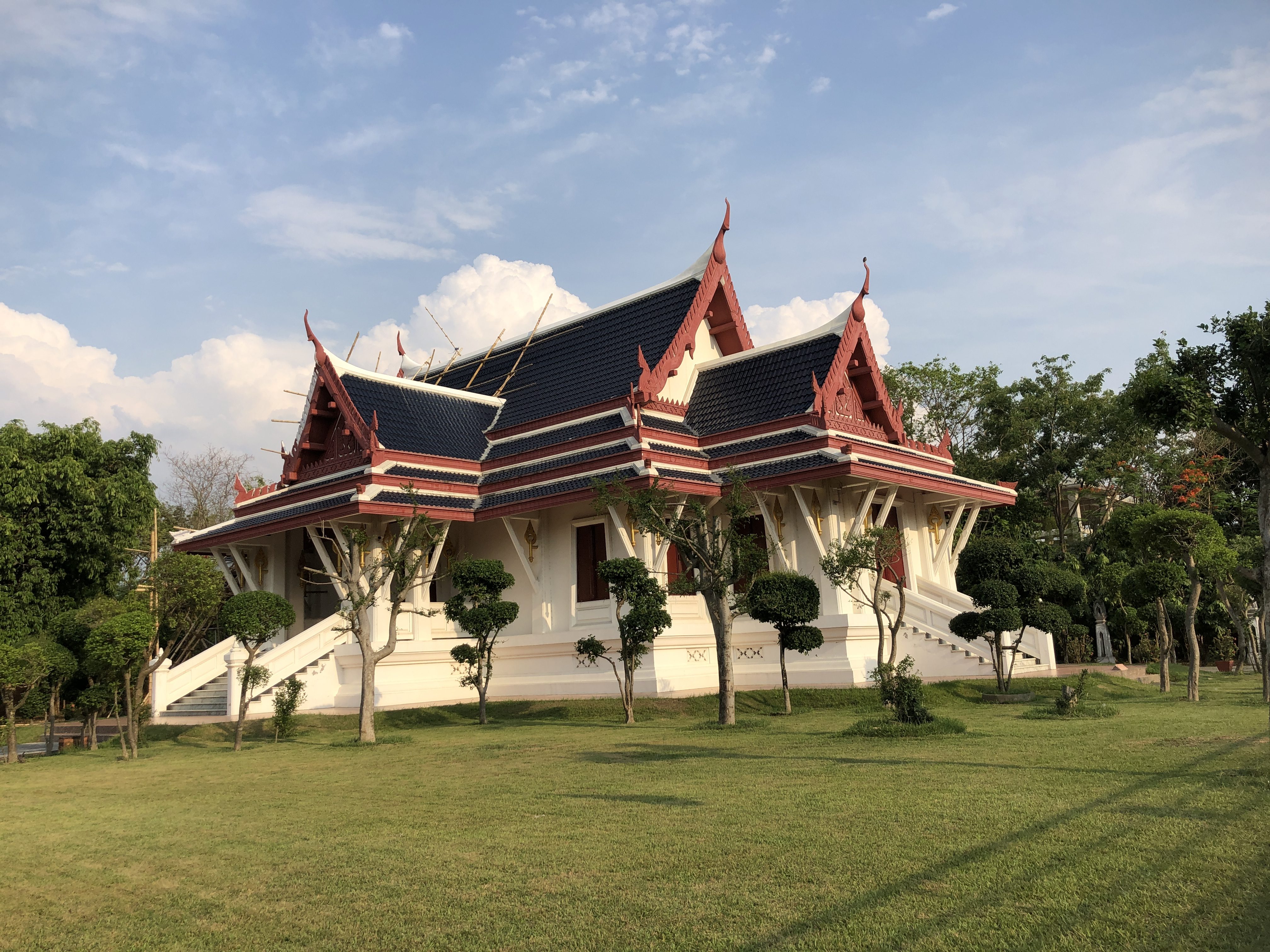 A traditional Thai-style building with ornate roof decorations and red and black tiles, surrounded by neatly trimmed trees and a grassy lawn under a blue sky with scattered clouds.