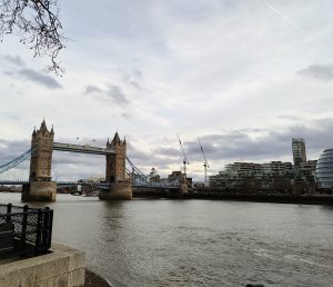 A view of Tower Bridge in London, spanning the River Thames under a cloudy sky, with modern buildings and construction cranes visible on the right