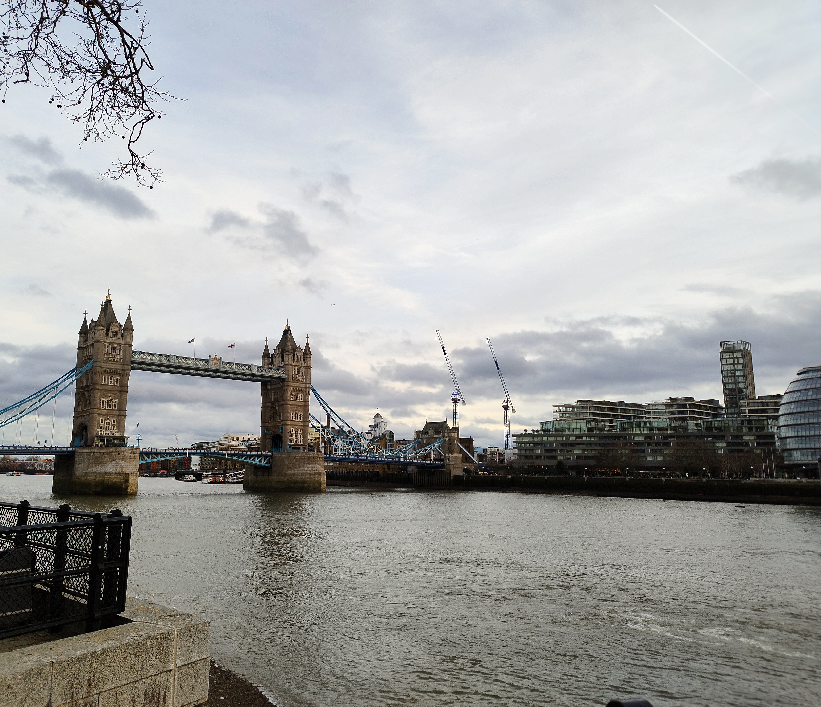 A view of Tower Bridge in London, spanning the River Thames under a cloudy sky, with modern buildings and construction cranes visible on the right