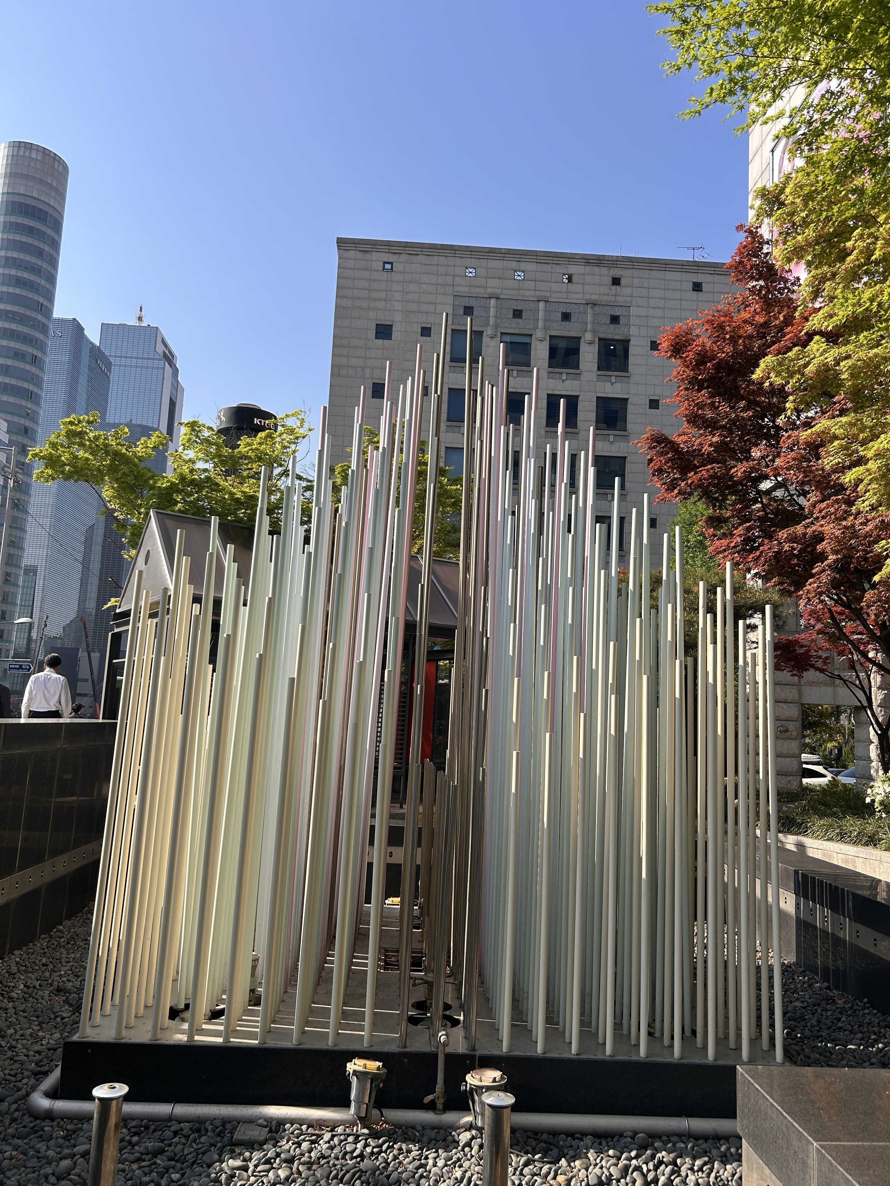 The image depicts an artistic installation made of tall, slender poles arranged closely together, creating a dense vertical structure