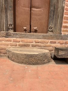A rustic, wooden door with visible age and wear, set into a brick wall. A stone step with a textured surface is in front, sitting on a red tiled floor. Small decorative carvings are on either side of the doorframe.