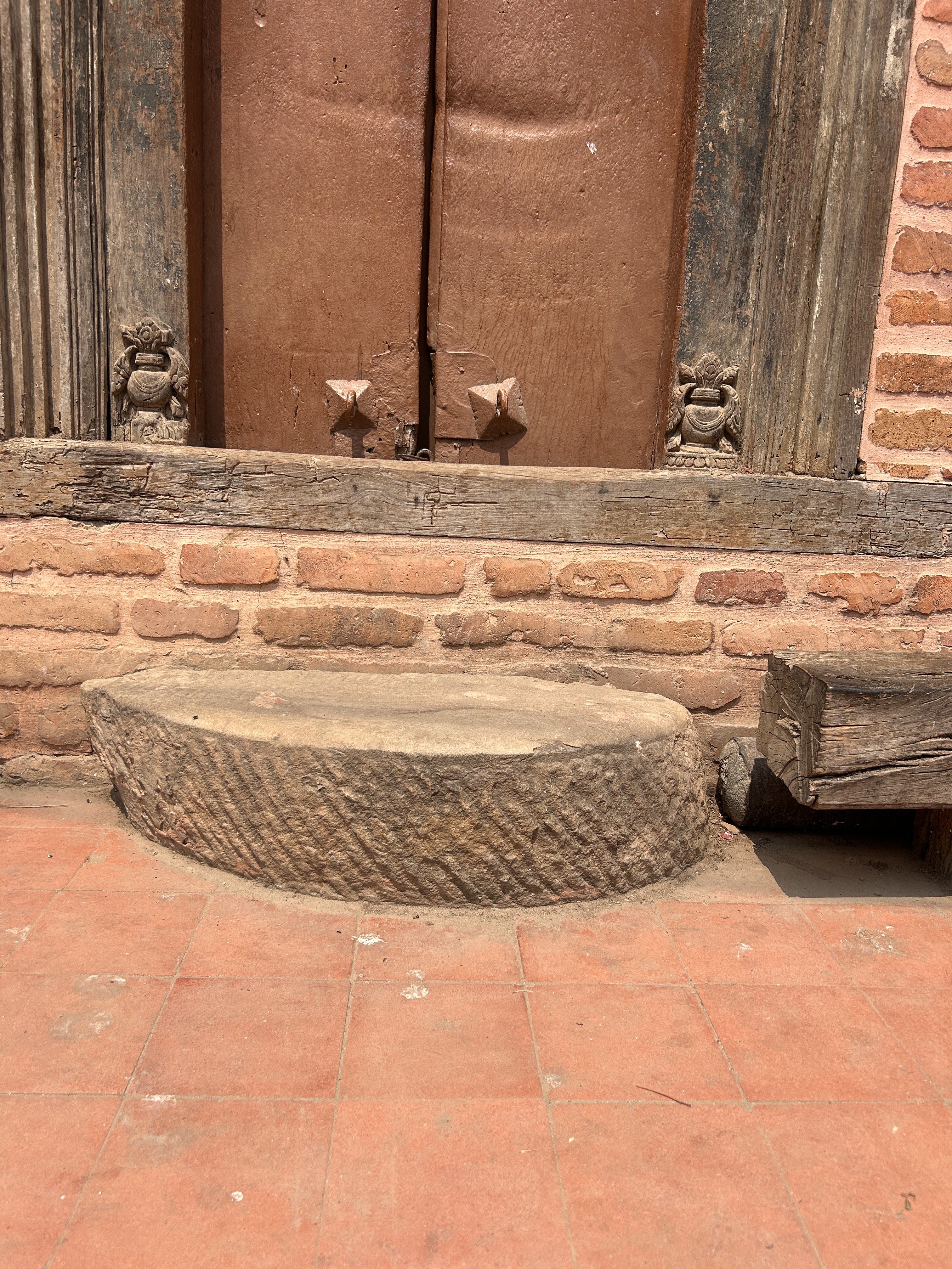 A rustic, wooden door with visible age and wear, set into a brick wall. A stone step with a textured surface is in front, sitting on a red tiled floor. Small decorative carvings are on either side of the doorframe.