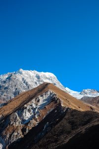 A triangular mountain peak with brown and rocky slopes in the foreground, partially covered with snow, set against a clear blue sky. In the background, larger snow-capped peaks are visible.