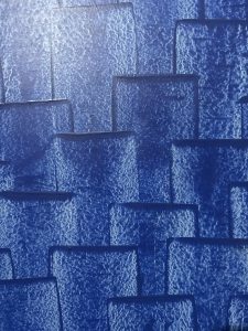 A textured blue surface with a pattern of overlapping, rectangular shapes creating a layered effect. The surface has a rough, slightly reflective finish.