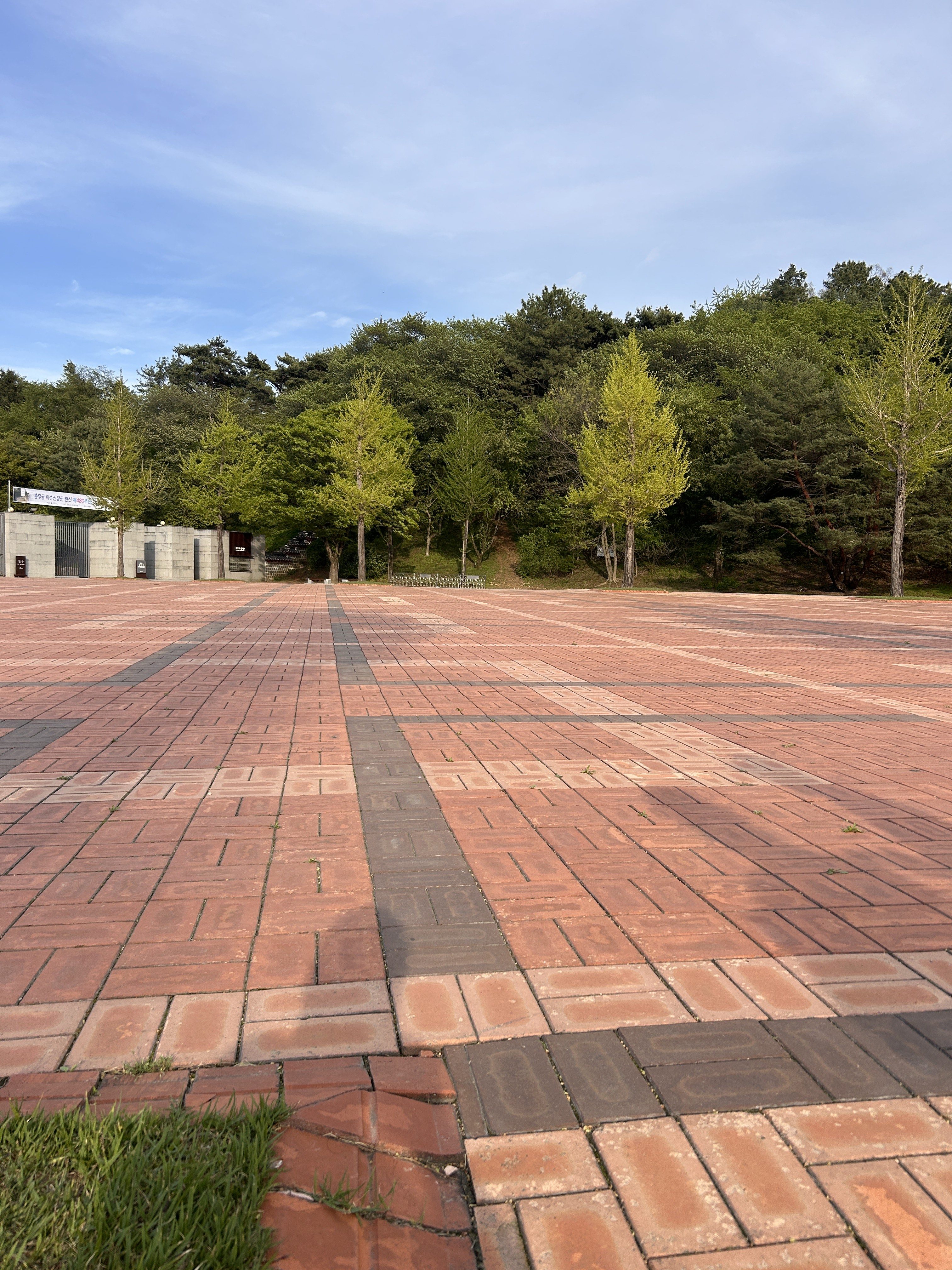 A wide, paved area made of red and black tiles stretches into the distance, bordered by lush greenery on one side. Young trees with bright green leaves are visible amidst the foliage, which leads up to a hill in the background.