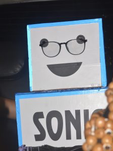 A boxy robot-like figure with a smiling face, black-rimmed glasses, and the word "SONIC" written on its torso, decorated with blue tape edges.