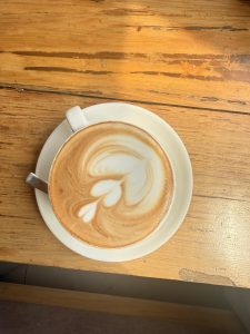 A cup of coffee with intricate latte art on top, placed on a saucer on a wooden table.