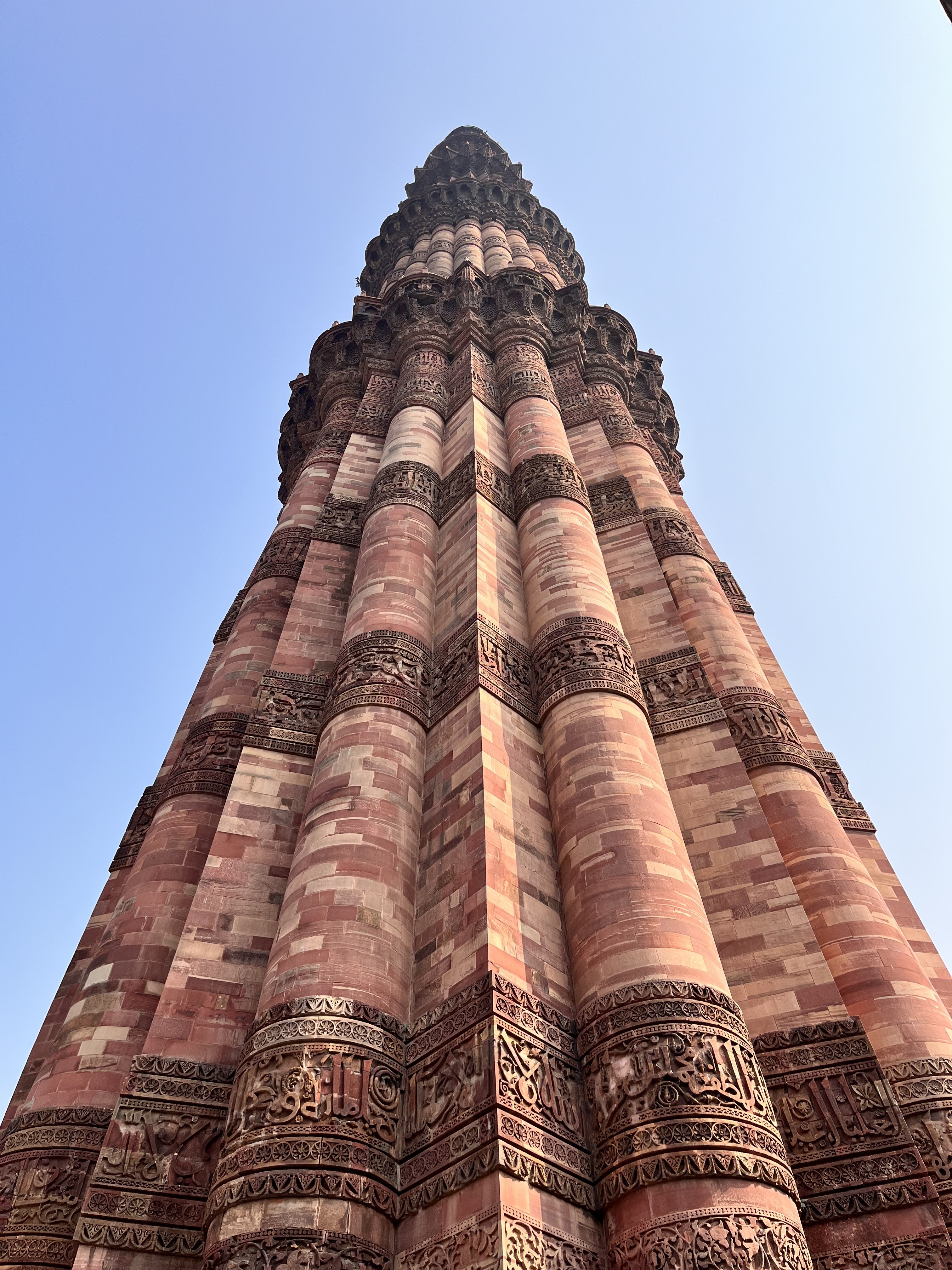 View looking up at a tall, intricately carved stone tower with reddish hues, featuring detailed carvings and inscriptions. The tower contrasts against a clear blue sky.